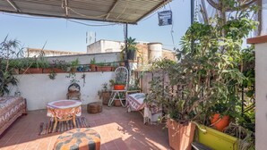 A rooftop patio with terracotta tiles, potted plants, a wooden table with a patterned tablecloth, a bench, and a chair.