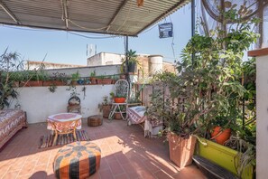 Une terrasse sur le toit, avec des carreaux de terre cuite, des plantes en pot, une table en bois recouverte d’une nappe à motifs, un banc et une chaise.