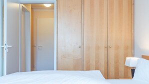 Quarto de hotel com uma cama, um guarda-roupa de madeira e uma mesa de cabeceira com uma luminária.