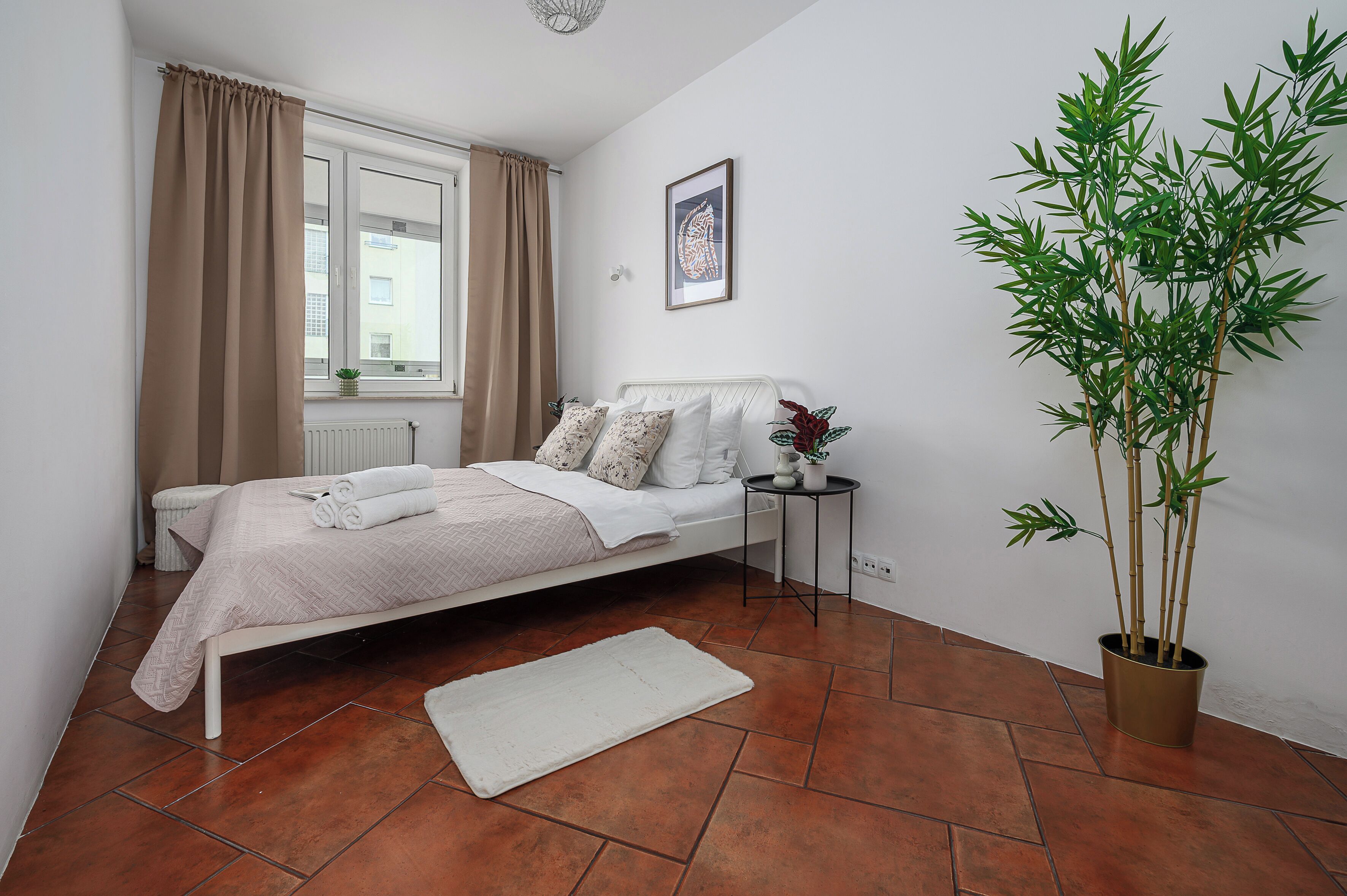 Un dormitorio con cama, mesita de noche, planta en maceta, radiador y ventana con cortinas.