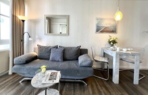 Ein modernes Wohnzimmer mit grauem Sofa, einem kleinen runden Tisch mit einer Zeitung, einem Beistelltisch mit Blumenvase, einem Schreibtisch mit Stuhl, einer Stehlampe, einem Spiegel und einem Wandbild.