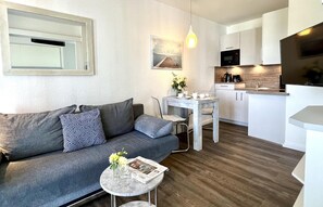 Un salon moderne avec un canapé gris, une petite table ronde sur laquelle est posé un magazine, et un coin repas avec une table et des chaises blanches.