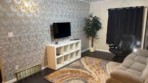 Un salon avec un téléviseur à écran plat, un meuble de rangement blanc, une grande plante en pot, un fauteuil en cuir noir et un canapé beige.