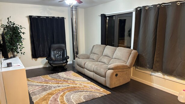 A living room with a sofa, a ceiling fan, a black chair, a large window with curtains, and a plant. - Cozy farmhouse retreat (Sherwood park)