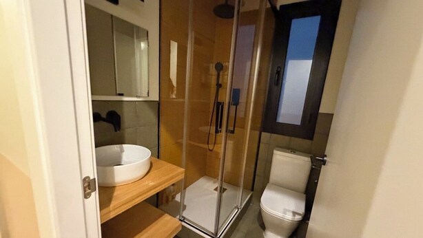 Un bany amb dutxa de vidre, un vàter blanc i un moble de tocador de fusta amb un lavabo blanc.