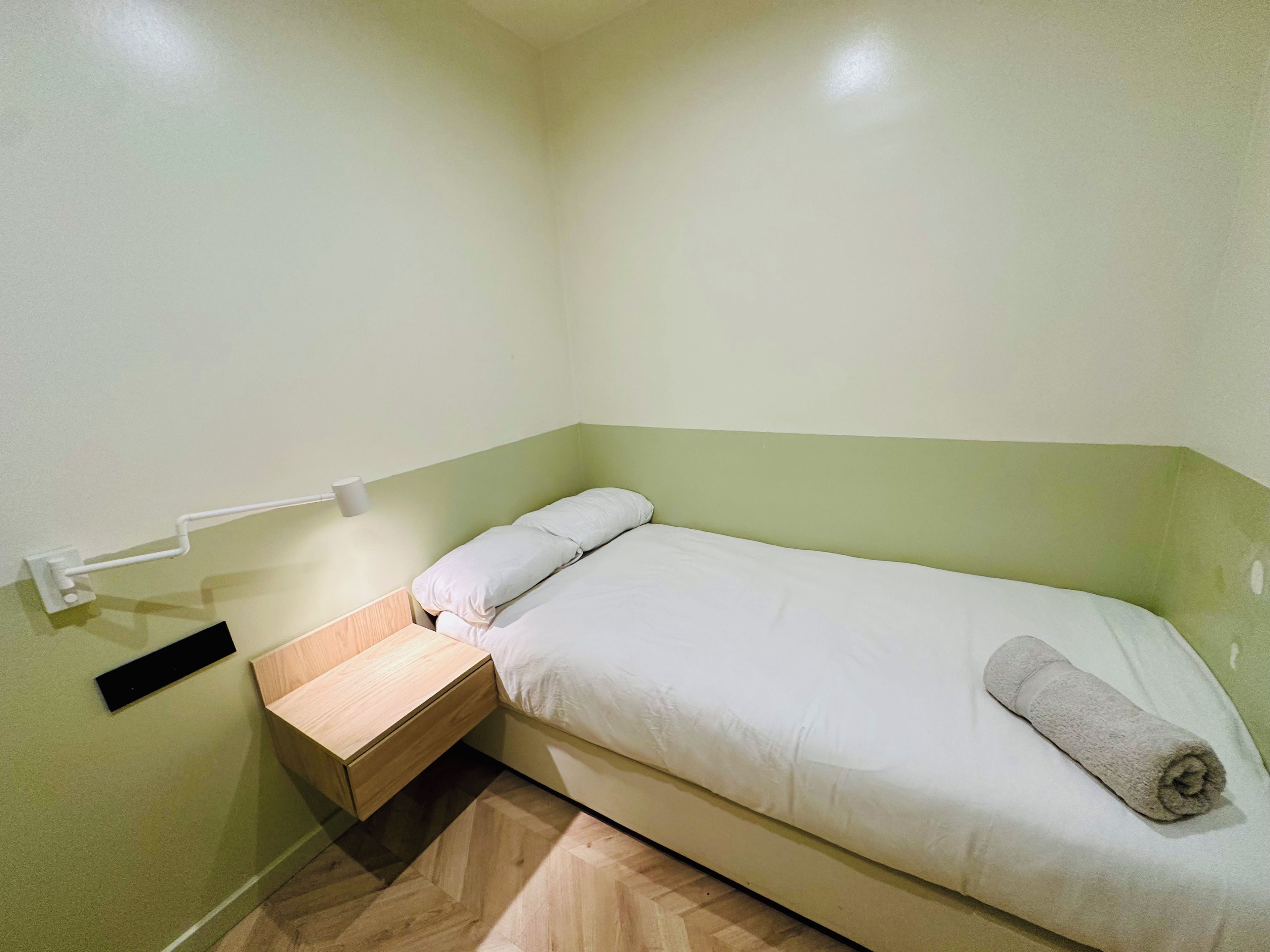 Ein kleines Zimmer mit einem Einzelbett, einem Holznachttisch, einem Handtuch auf dem Bett und einer Wandleuchte.