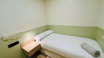 Ein kleines Zimmer mit einem Einzelbett, einem Holznachttisch, einem Handtuch auf dem Bett und einer Wandleuchte.
