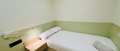 Une petite chambre avec un lit simple, une table de chevet en bois, une serviette sur le lit et un luminaire fixé au mur.