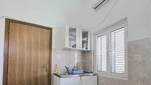 Una piccola cucina con una porta in legno, mobili bianchi, una finestra con persiane e un condizionatore d'aria.