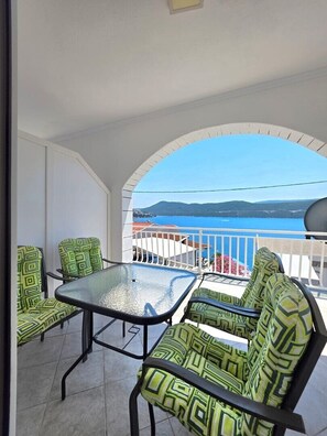 Ein Balkon mit Glastisch und Stühlen, einem weißen Geländer und Blick auf das Meer und die Hügel.