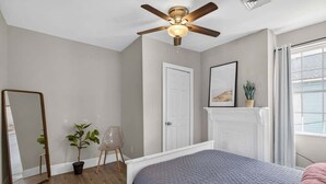 Ein Schlafzimmer mit einem Bett, Deckenventilator, einer Tür, einem Spiegel, einer Pflanze und einem Fenster.