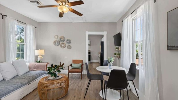 Ein modernes Wohnzimmer mit Deckenventilator, einer Couch, einem Esstisch mit Stühlen und einem an der Wand befestigten Fernseher.