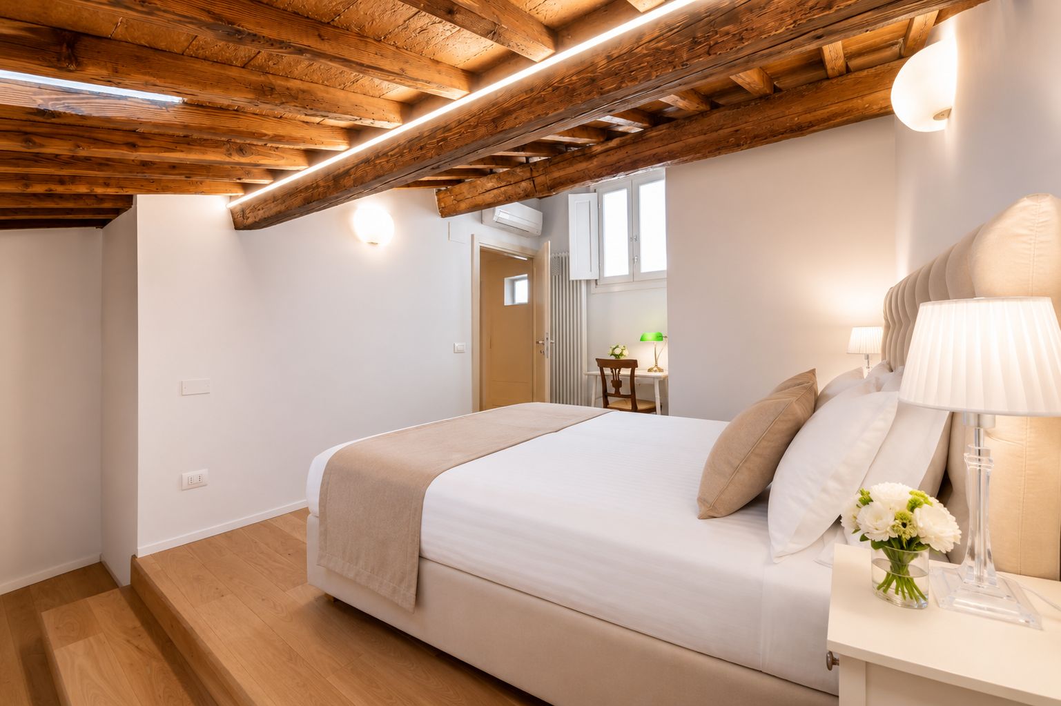 Un dormitorio con techo de madera, una cama con ropa blanca, una mesita de noche con lámpara y un florero con flores.