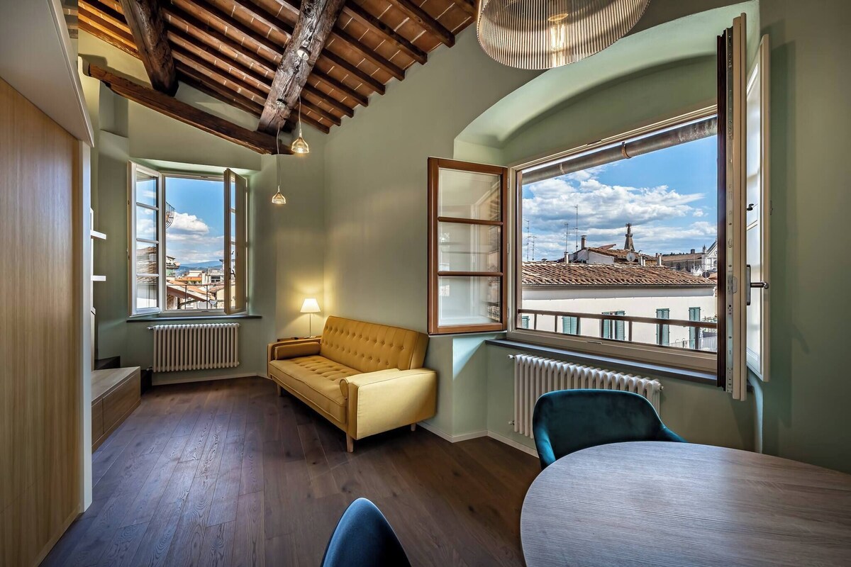 A room with a wooden ceiling, a sofa, a table, and a view of a cityscape through large windows.