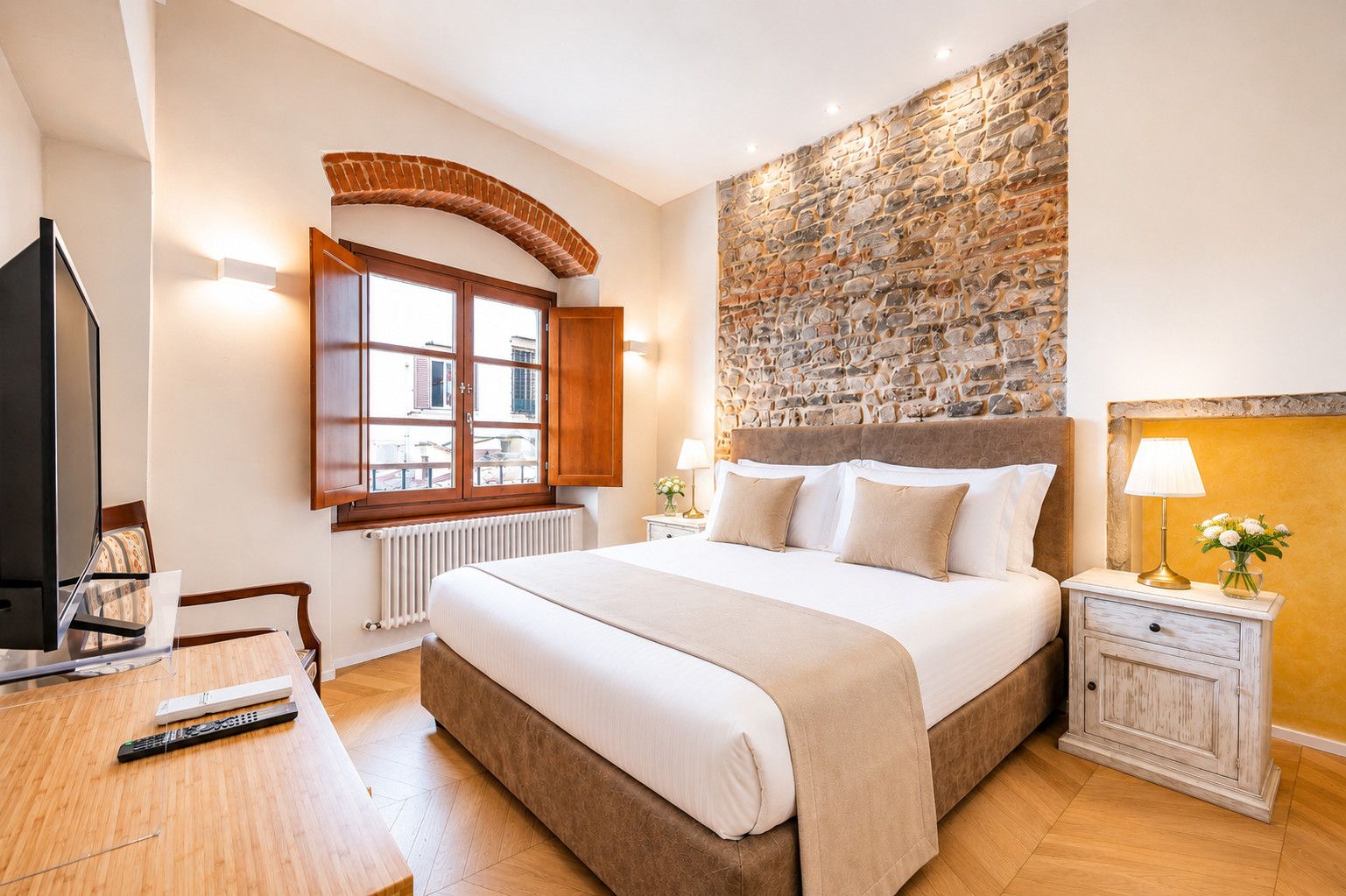 Habitación de hotel con una cama grande, un escritorio de madera con televisor, una pared de piedra y una ventana con vistas a edificios.