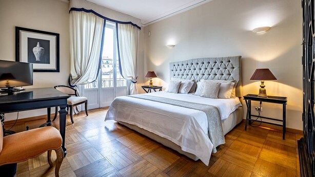 Ein Schlafzimmer mit einem großen Bett, einem Schreibtisch, zwei Nachttischen, einem Sessel und einem Fenster mit Vorhängen.