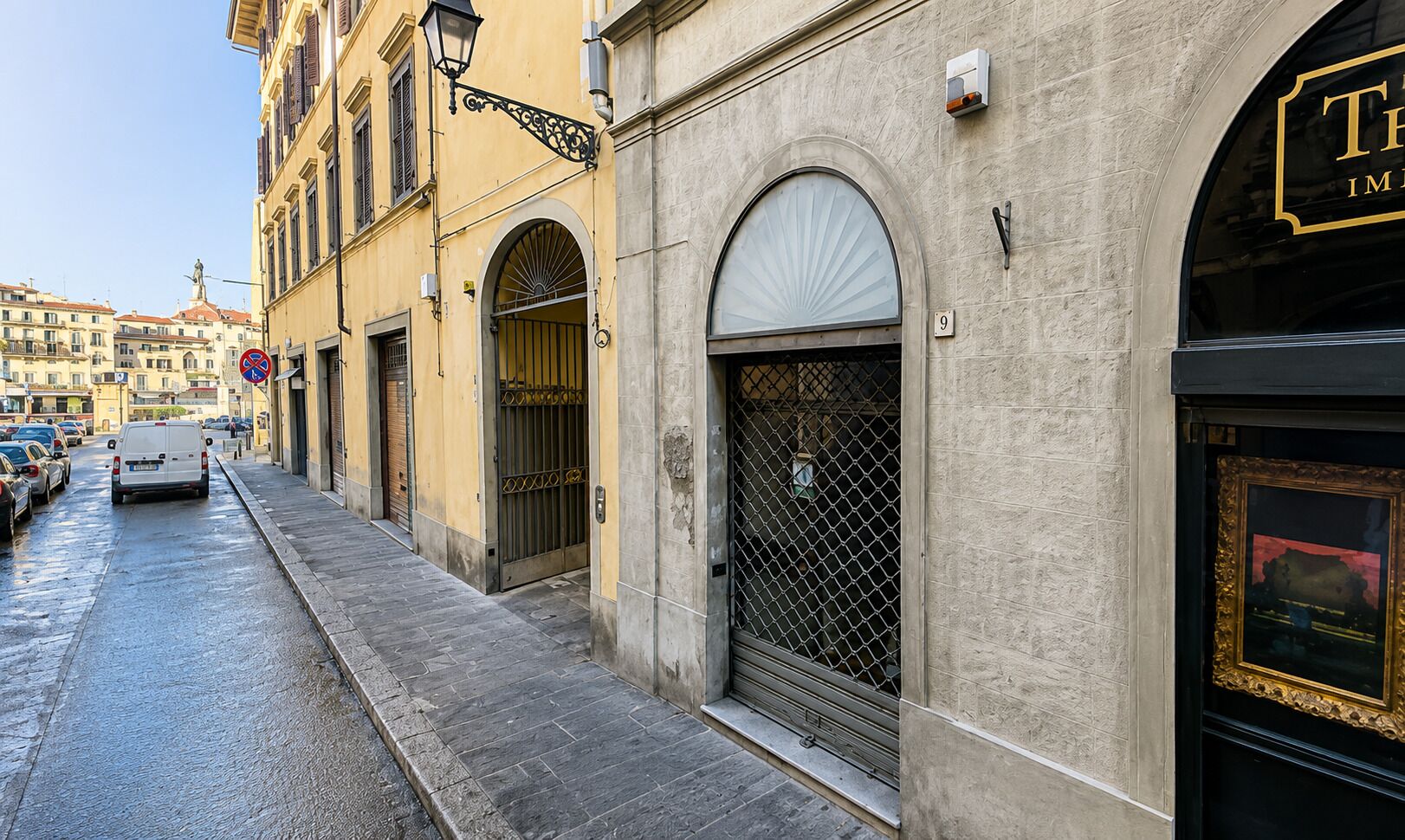 A street view with a building featuring a large arched entrance, a visible number '9' sign, and a black-framed window with a gold frame.