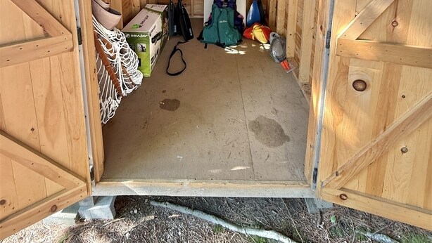 Un capanno di legno con tetto spiovente, una piccola finestra e uno sfiato sul tetto. All'interno, due pagaie, uno zaino verde e una borsa bianca.