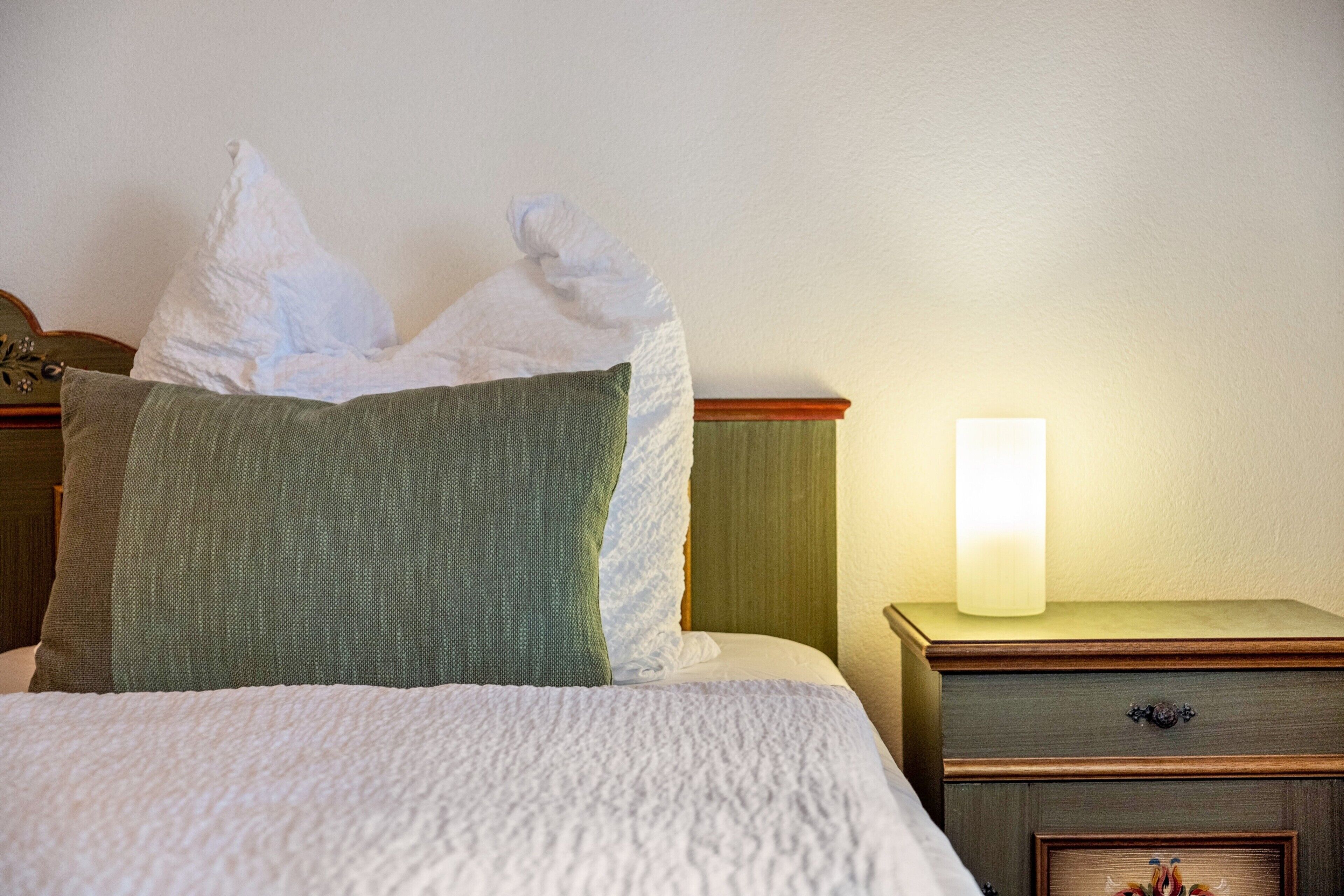 Ein ordentlich bezogenes Bett mit einer weißen, strukturierten Decke, einem grünen Kissen und einem weißen Kissen. Ein Holznachttisch mit einer Lampe und einer Schublade mit einer Dekoration.