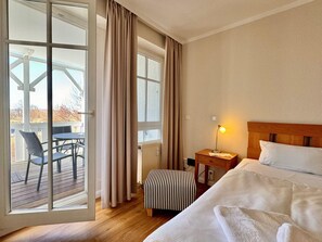 Ein Hotelzimmer mit einem Bett, einem Nachttisch mit Lampe, einem gestreiften Hocker, einem Fenster mit Vorhängen und Blick auf eine Terrasse mit Stühlen.