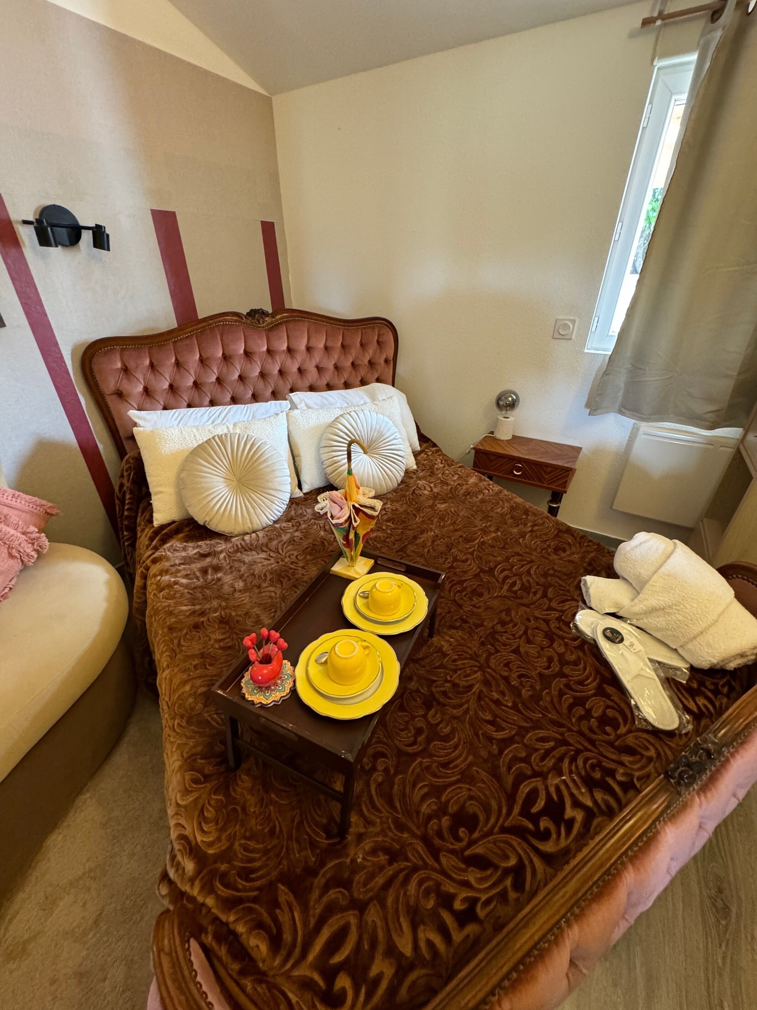 A bedroom with a tufted headboard, a bed with patterned bedding, a bedside table with a lamp, and a window with curtains.