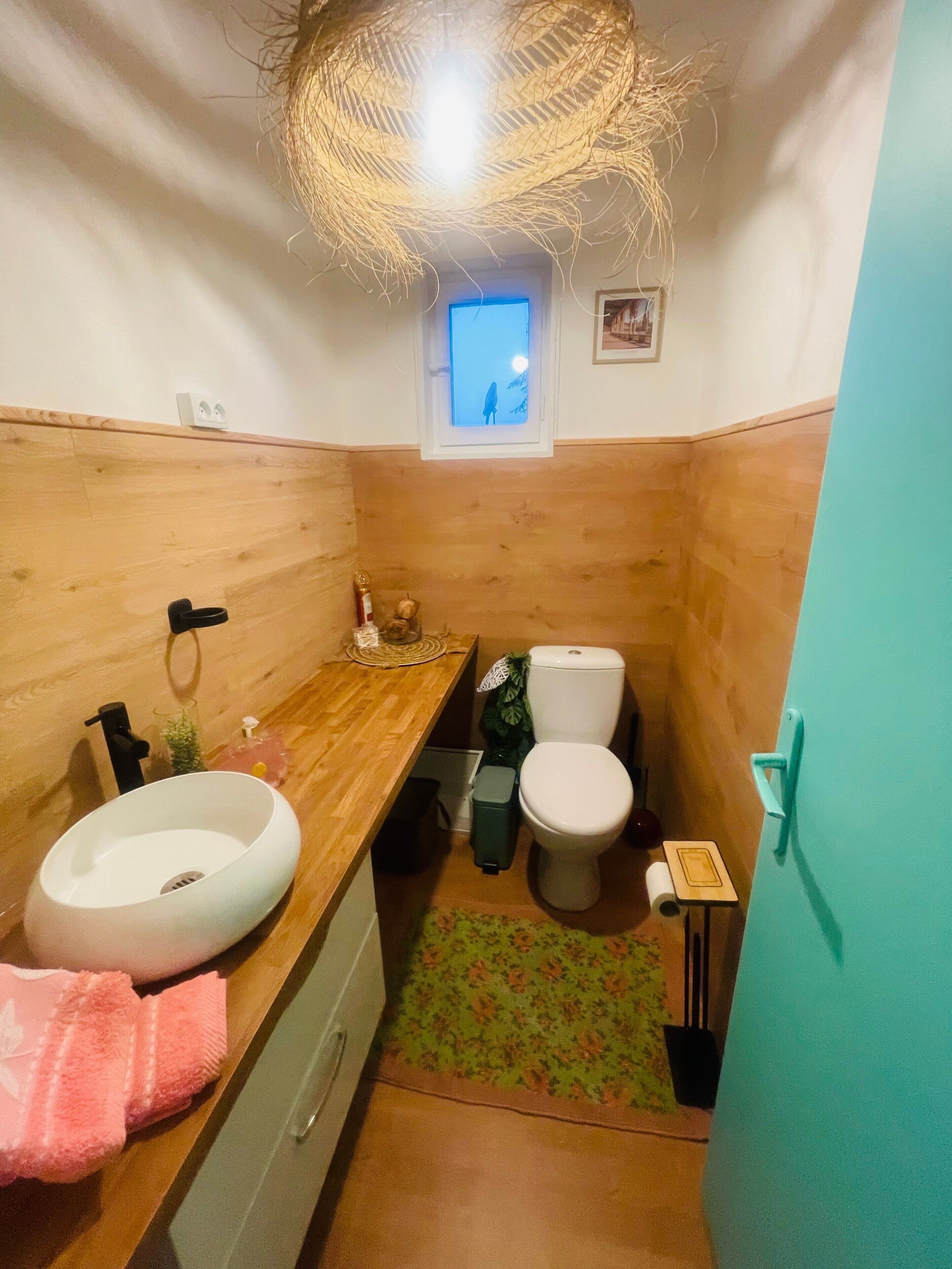 A compact bathroom with a wooden vanity, a white sink, a toilet, a small window, and a green floral rug.