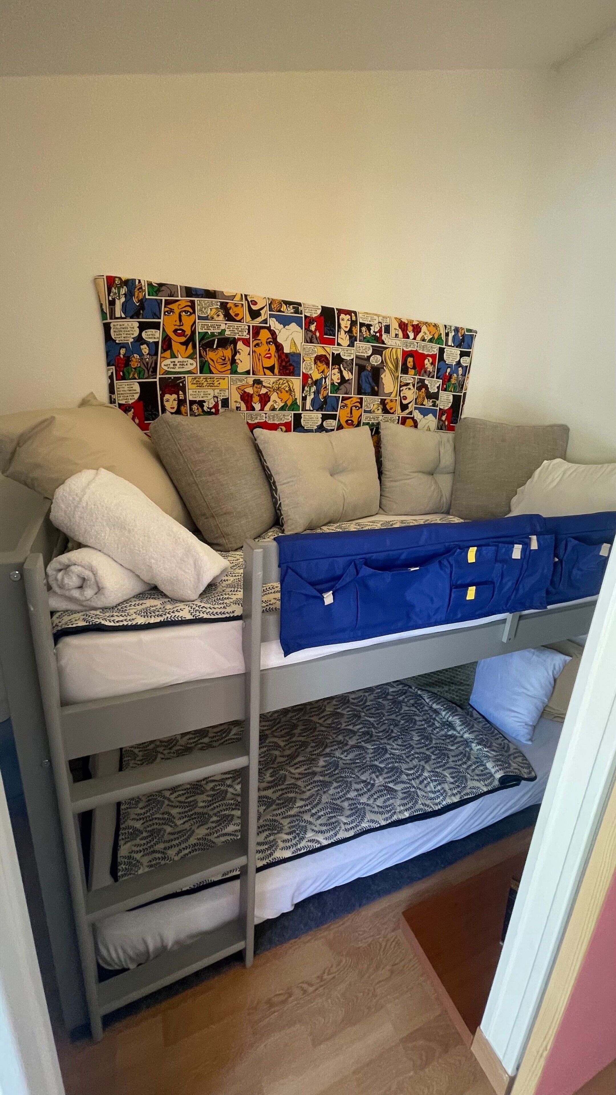 Een stapelbed met stripboek decoratie aan de muur, een grijs hoofdbord en een getextureerd vloerkleed.