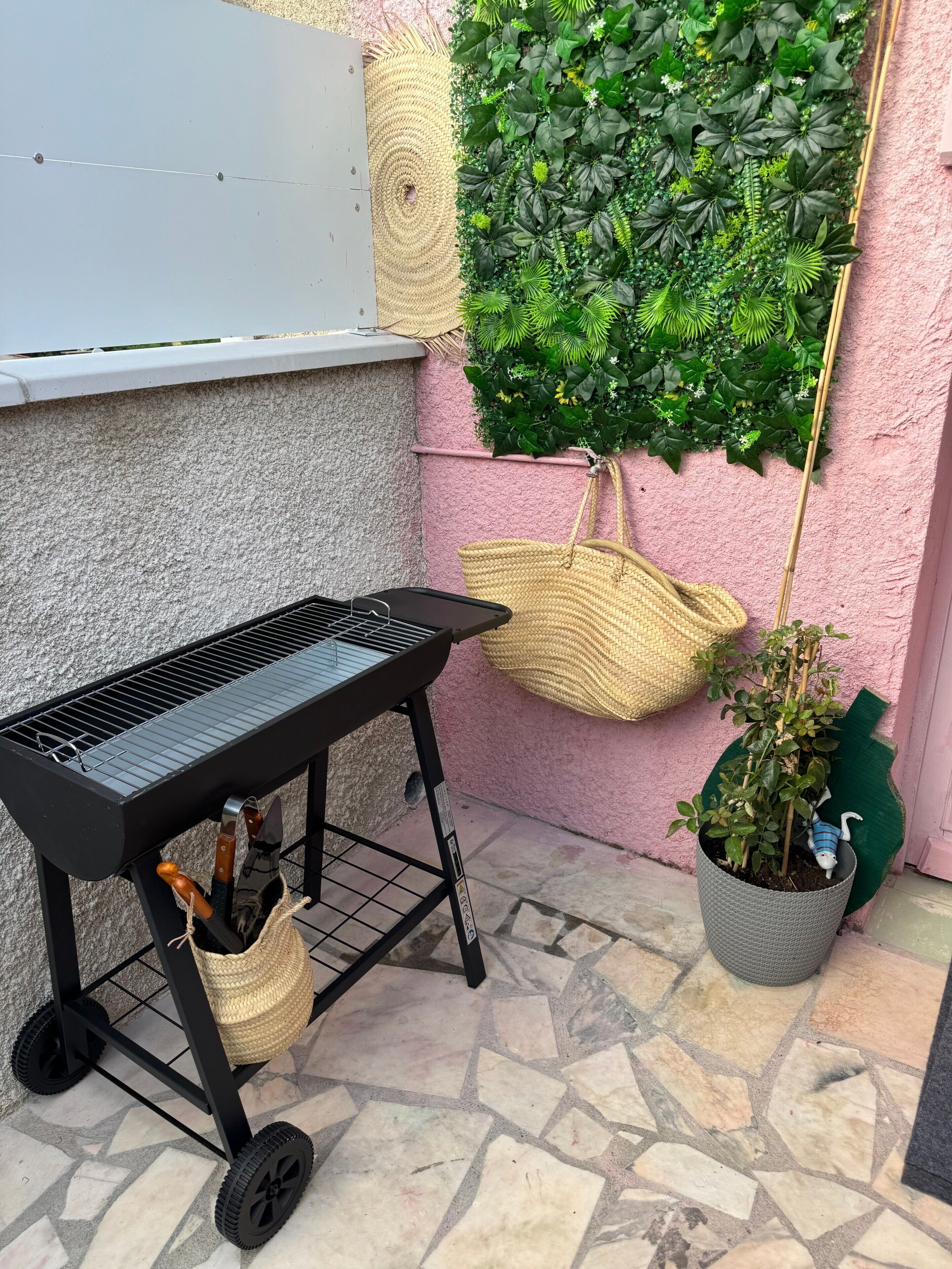 Ein Grill mit Metallrost und Rollen, ein gewebter Korb und ein Blumentopf mit einer Spielzeugfigur.