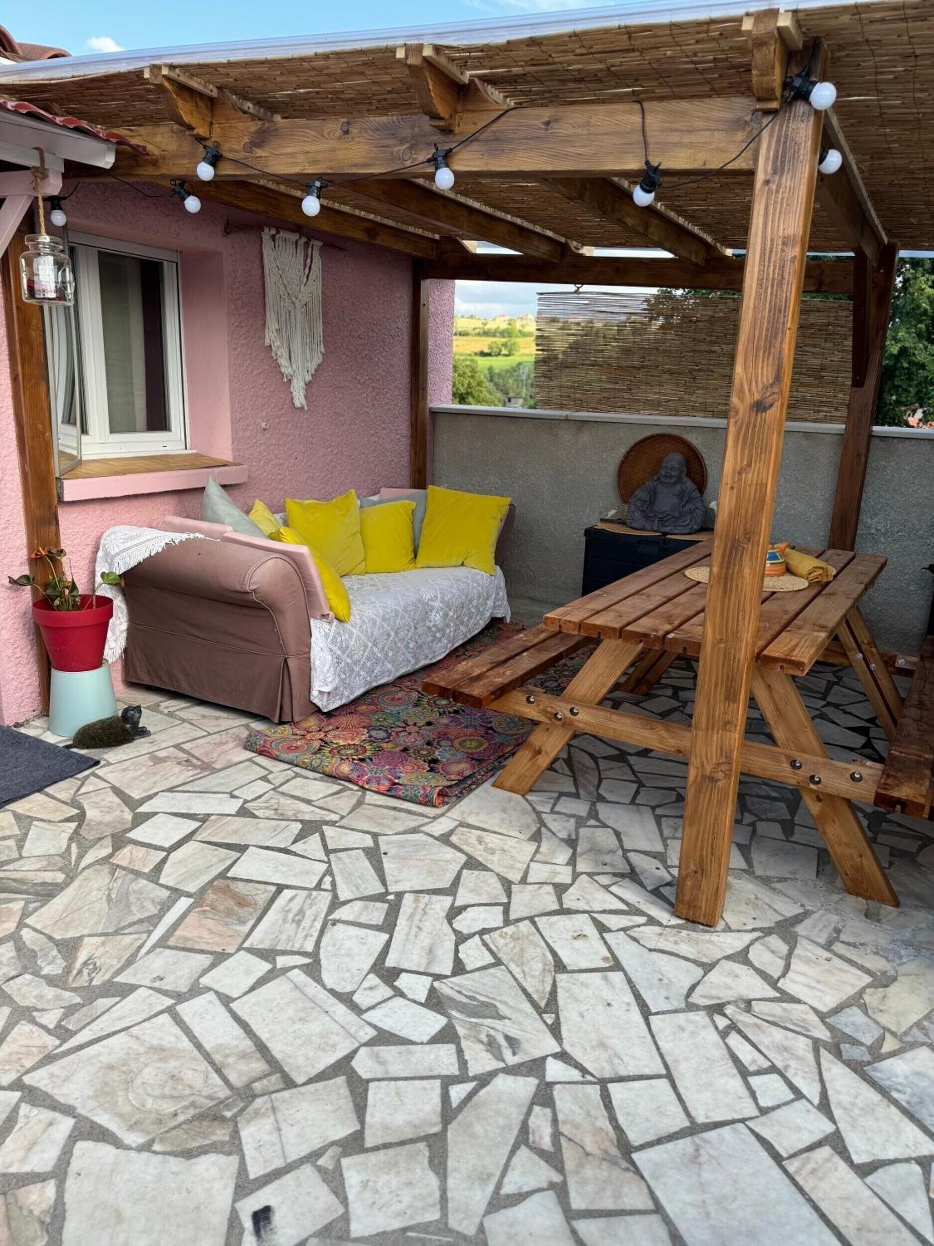 Un espace de détente extérieur accueillant, avec un banc en bois, un banc rembourré avec des coussins jaunes et un tapis à motifs.