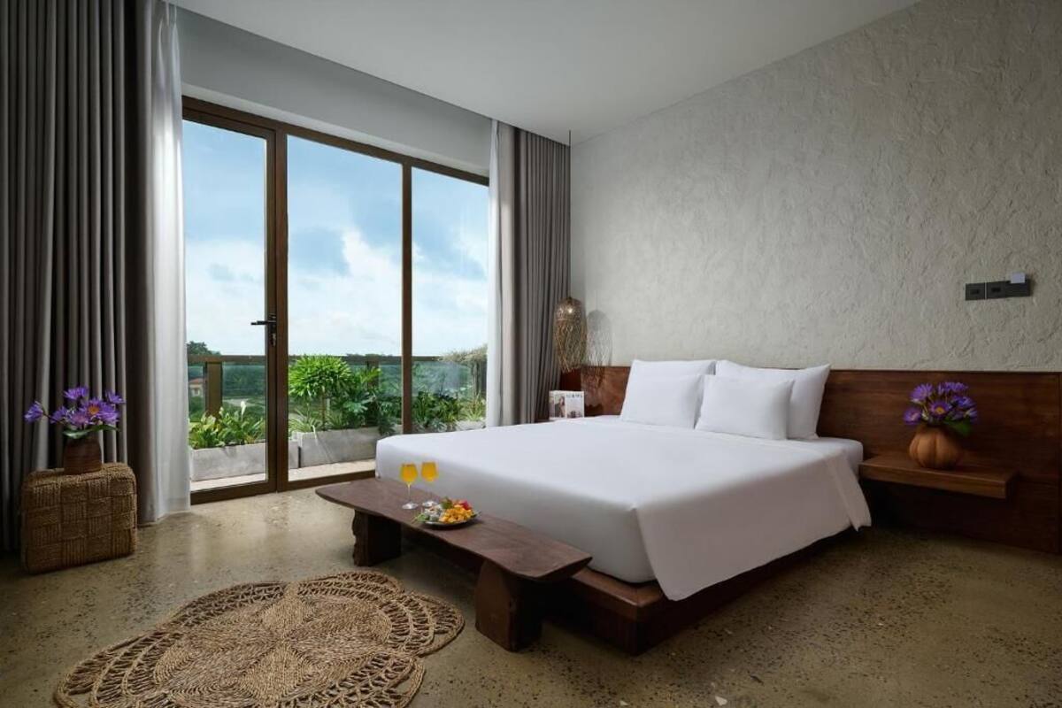 Une chambre d’hôtel moderne avec un grand lit, un banc en bois, un tapis tissé et une vue sur la verdure par la fenêtre.