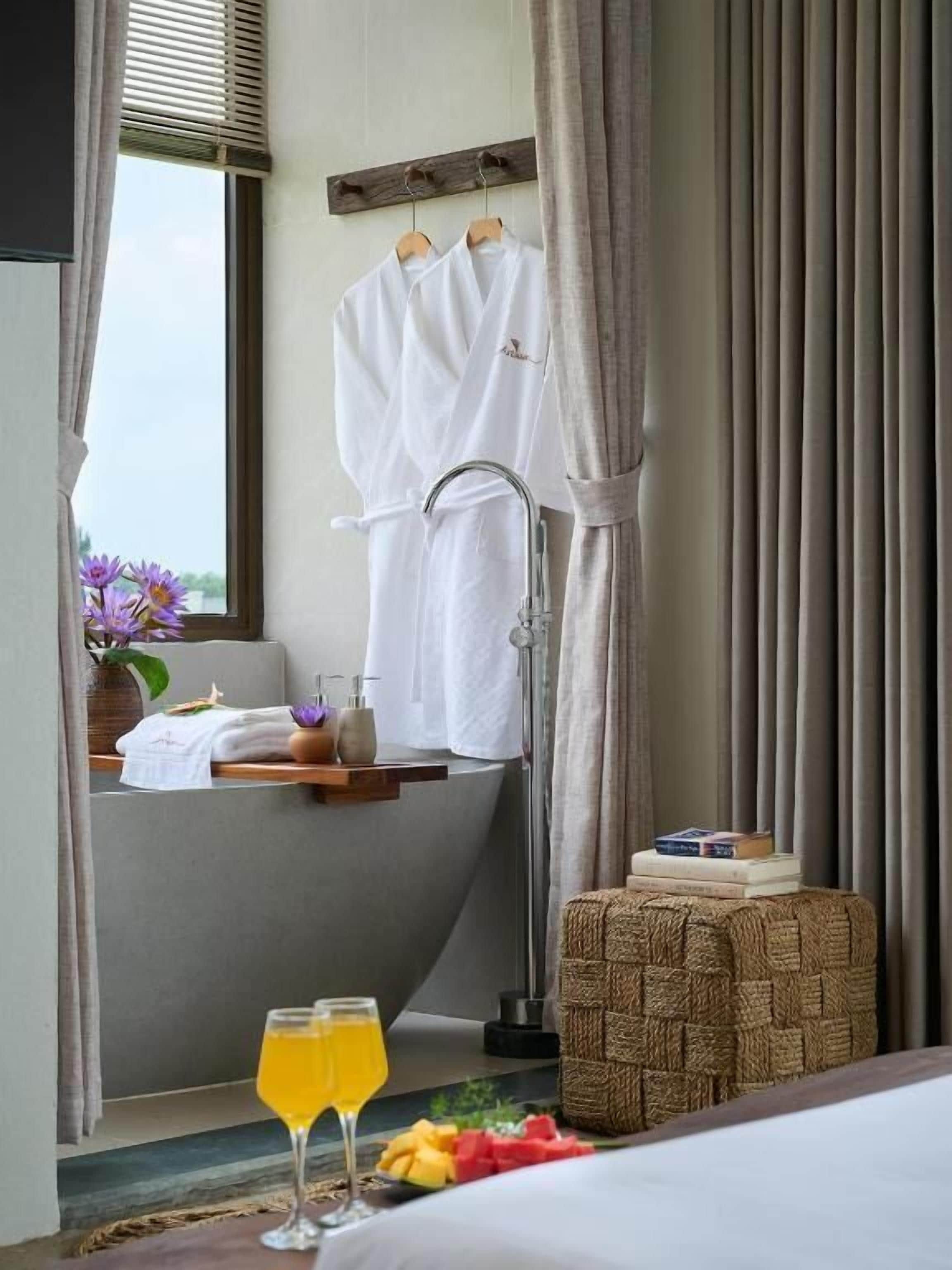 A hotel room with a bathtub, a vanity with a mirror, a bedside table with books and a drink, and a window with curtains.