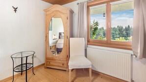 Una habitación con techo de madera, una ventana con cortinas, un radiador blanco, un armario de madera con espejo, una silla blanca y una mesa de vidrio.