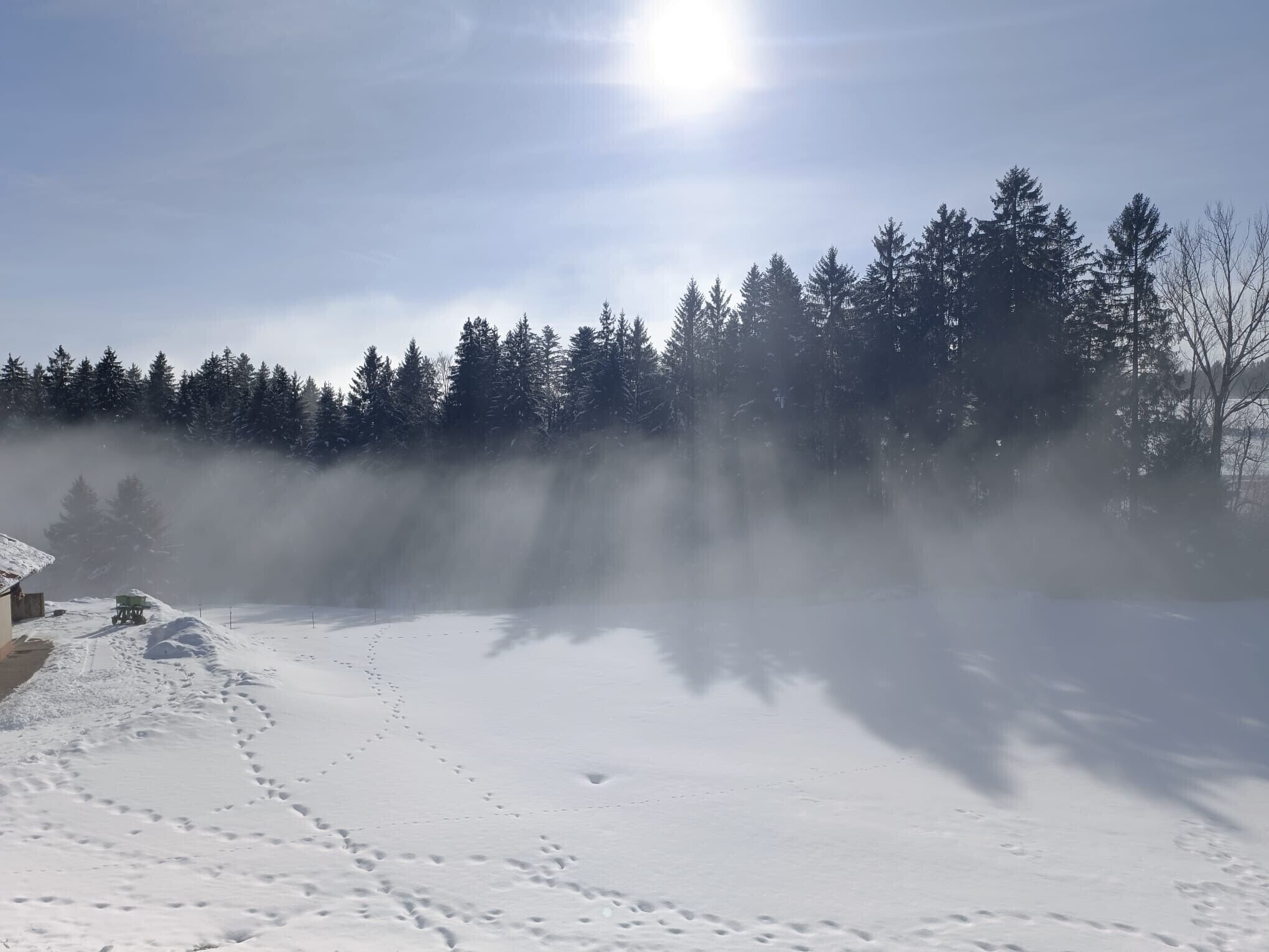 Schneeverdeckte Landschaft mit blauem Himmel, einem Pfad und einem Gebäude, das teilweise im Nebel verborgen ist.