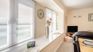 Una sala de estar con sofá, televisor, ventana con persianas, un reloj y un florero con flores.