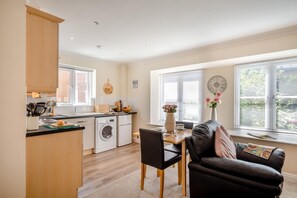A compact kitchen with a washing machine, a dining table with chairs, and a sofa in a bright room with large windows. - 1 Bed in Whitby (oc-w815) (Whitby)