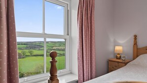 Un dormitorio con un ventanal grande, una cama de madera, una mesita de noche con una lámpara y vistas a campos verdes.