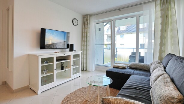 En moderne stue med sofa, sofabord i glas, et hvidt skab med glaslåger, et fladskærms-tv og udsigt til en balkon med bypanorama.