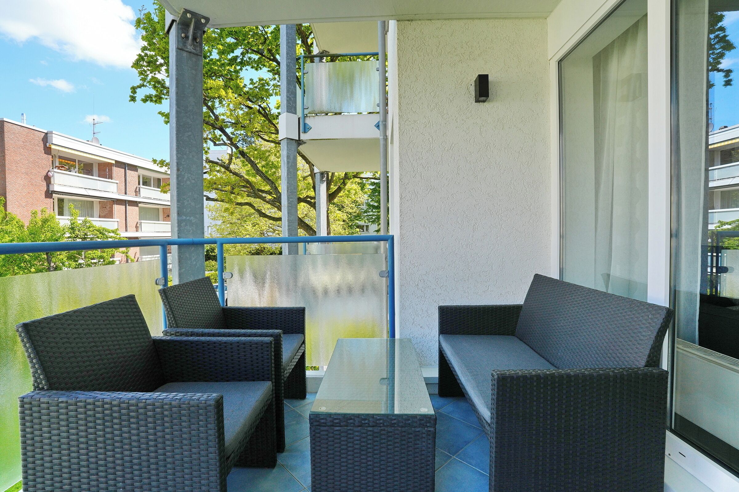 Un balcon agrémenté de meubles en osier, d’une table en verre et offrant une vue sur les immeubles et les arbres.