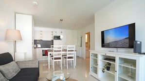 En moderne stue med en sofa, et glasbord, et spiseområde med stole, et køkken i baggrunden og et fjernsyn ophængt på væggen.