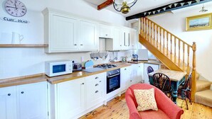 A kitchen with white cabinets, a microwave, and a stove. There's a red chair and a wooden staircase. - 1 Bed in Whitby (oc-r049) (Robin Hood's Bay)