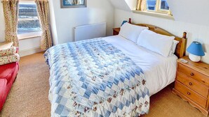 A bedroom with a bed, a nightstand, a red chair, a window with curtains, and a framed picture on the wall. - 1 Bed in Whitby (oc-r049) (Robin Hood's Bay)