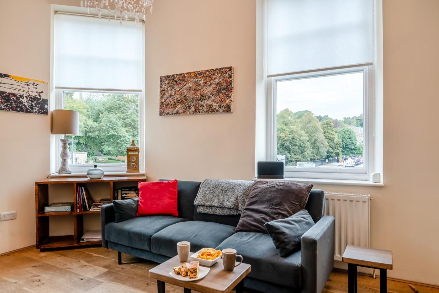 Ein modernes Wohnzimmer mit einer dunkelgrauen Couch, einem kleinen Couchtisch und einem Bücherregal. Es gibt zwei Fenster mit Jalousien, eine Pendelleuchte und ein Bild an der Wand.