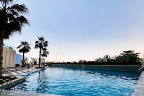 Ein Schwimmbecken mit Liegestühlen und Palmen, ein Riesenrad im Hintergrund und ein modernes Gebäude.