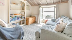 A cozy living room with a sofa, a bookshelf, a wooden chest, and a view through a window. - 2 Bed in Looe (oc-l10094) (Looe)