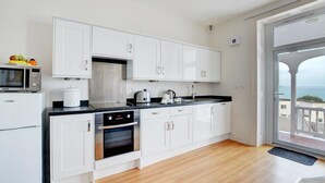 A modern kitchen with white cabinets, a black countertop, a microwave, an oven, and a view of the sea through a balcony. - 1 Bed in Looe (oc-l30006) (Looe)