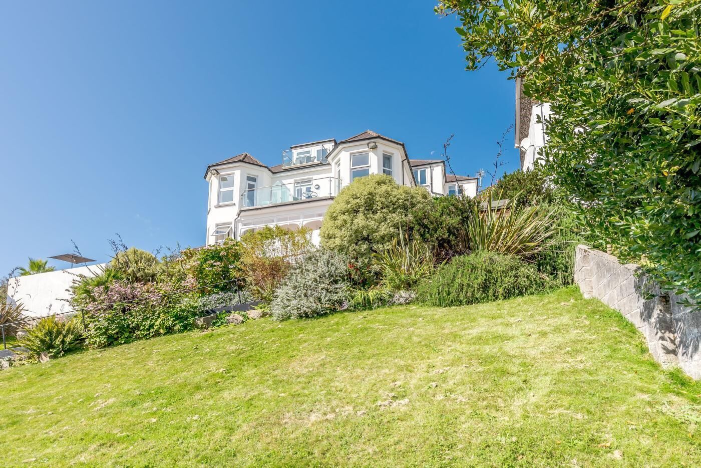 A large, multi-story house with a prominent balcony, surrounded by lush greenery and a sloped lawn.
