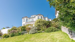 A large, multi-story house with a prominent balcony, surrounded by lush greenery and a sloped lawn. - 1 Bed in Looe (oc-l30006) (Looe)
