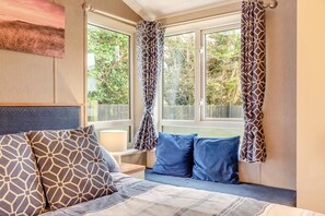 Ein Schlafzimmer mit Bett, Kissen, Nachttisch, Lampe und einem Fenster mit Vorhängen.