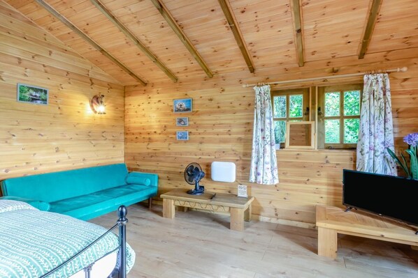 A wooden cabin interior with a turquoise sofa, a television, a fan, and a window with curtains. - 1 Bed in Westley Waterless (oc-b34150) (Westley Waterless)