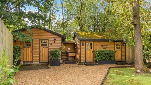 A wooden cabin with a gravel driveway, a wooden fence, and a tree.
